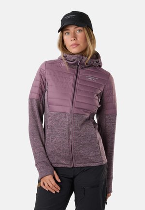 Kobieta nosząca jasnoróżową kurtkę z kapturem z pikowanymi górnymi panelami i czarną czapką, stojąca na tle jednolitego białego tła.