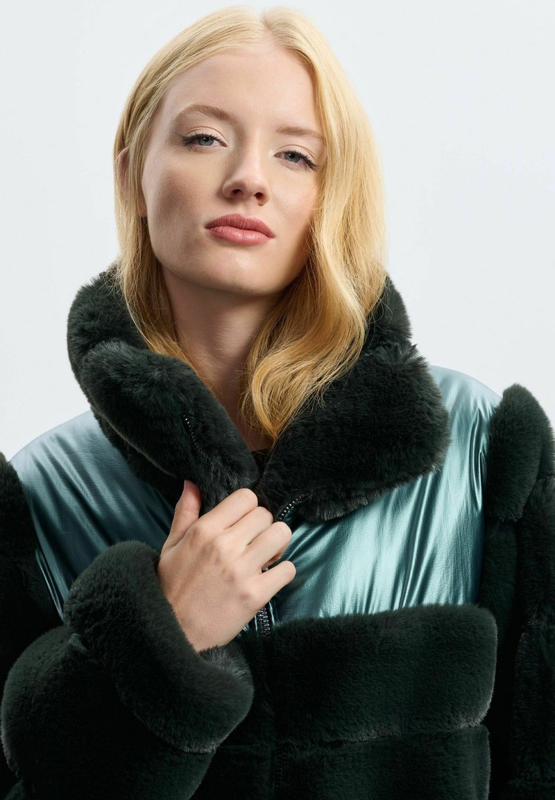 Cappotto in pelliccia artificiale con una base verde scuro e dettagli lucidi in teal, caratterizzato da un colletto alto, chiusura con zip e una texture morbida.