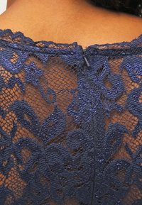 Tissu en dentelle marine avec des motifs floraux, présentant une texture délicate. Comprend une fermeture éclair alignée verticalement à l'arrière.