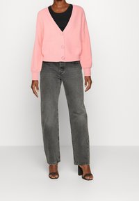 Rosa gestrickte Strickjacke mit V-Ausschnitt und Knopfverschluss, kombiniert mit grauen Straight-Leg-Jeans und schwarzen Sandalen mit Absatz.