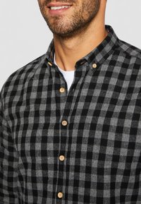 Camicia in flanella a quadri neri e grigi con colletto a bottone, caratterizzata da bottoni in legno e una texture morbida.