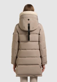 Puffa kabát v taupe barvě s kapucí z umělé ovčí vlny, s vodorovným prošíváním a černým táhlem zipu na zadní části.
