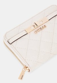 Portafoglio beige trapuntato con hardware dorato e chiusura con zip, caratterizzato da un accento logo e materiale testurizzato. Forma rettangolare compatta.