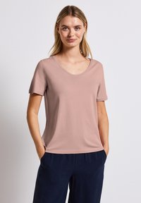 Světle růžové tričko s krátkým rukávem a kulatým výstřihem, které má hladkou texturu a volný střih, v kombinaci s tmavě námořnickými kalhotami.