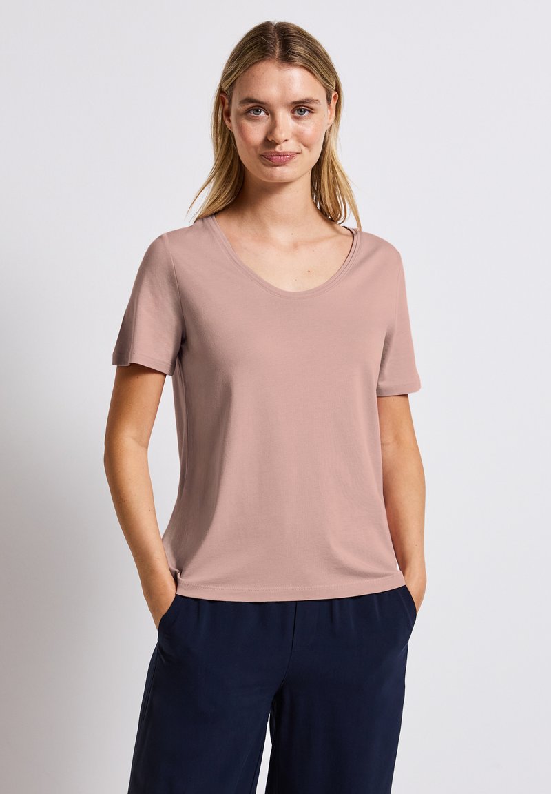 Světle růžové tričko s krátkým rukávem a kulatým výstřihem, které má hladkou texturu a volný střih, v kombinaci s tmavě námořnickými kalhotami.