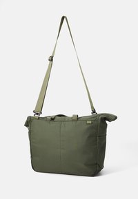 Sac fourre-tout en tissu vert de forme rectangulaire, avec une bandoulière ajustable et des poignées supérieures. Il possède un rabat en haut et un design minimaliste.