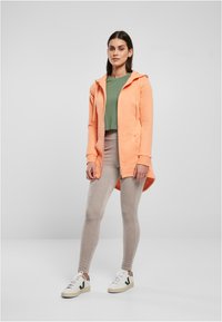 Ružová mikina na zips so širokým golierom, v kombinácii so zeleným crop topom a béžovými legínami. Modelka má na sebe biele tenisky so zelenými akcentmi.