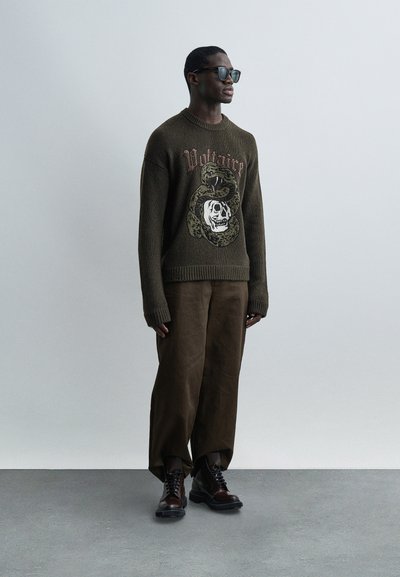 Άνδρας με σκούρα γυαλιά ηλίου, καφέ πουλόβερ με σχέδιο φίδι και κρανίο, φαρδιά καφέ παντελόνια και σκούρα καφέ μπότες, στέκεται μπροστά σε ανοιχτό γκρι φόντο.