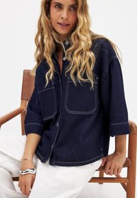 Kvinde med langt bølget blond hår, siddende på en træstol, iført en mørkeblå denimskjorte med hvid stikning og hvide bukser.