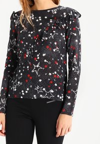 Long-sleeve top in black fabric with star pattern in red, white, and gray. Features ruffle accents along shoulders and a relaxed fit.