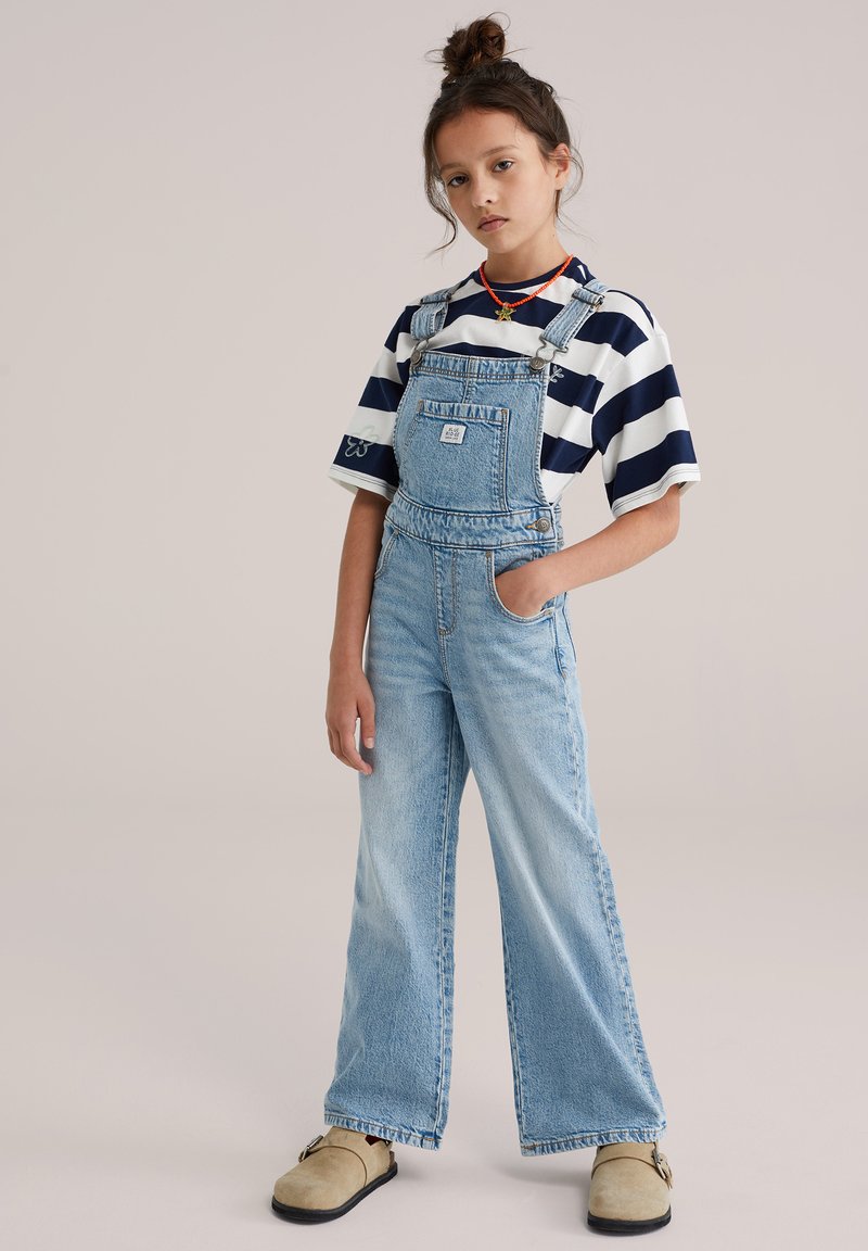 Salopetă din denim albastru deschis, cu croială cu picioare largi, bretele reglabile, buzunar frontal, asortată cu un tricou în dungi bleumarin și alb și saboți bej.