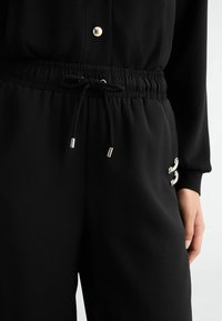 Čierne jogger nohavice s elastickým pásom, detail sťahovacej šnúrky a kovovými akcentmi na boku, majú hladkú textúru a voľný strih.