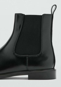 Bottine Chelsea noire avec une tige en cuir brillant, des panneaux élastiques sur les côtés et une semelle en caoutchouc plate. Reconnaissable par sa silhouette élégante et son design minimaliste.