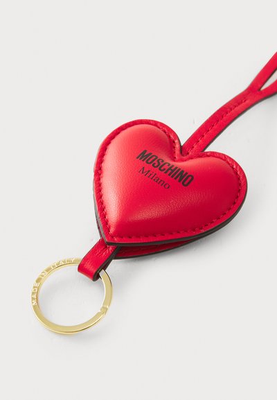 Breloc din piele roșie, în formă de inimă, cu inel aurit. Prezintă logo-ul "Moschino Milano" pe partea din față și o suprafață texturată.