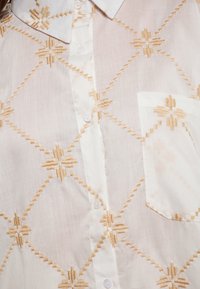 Witte blouse met verfijnde gouden borduursels in een ruitpatroon, voorzien van een puntige kraag, knoopsluiting en een linkse borstzak.