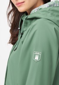 Grön vattentät jacka med en jämn yta, justerbar huva och dekorativ paisleymönstrad foder. Har ett logopatch på axeln.