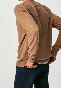 Personne portant un pull marron à manches longues et un jean bleu foncé, ajustant l'arrière du pull avec les mains près des hanches.