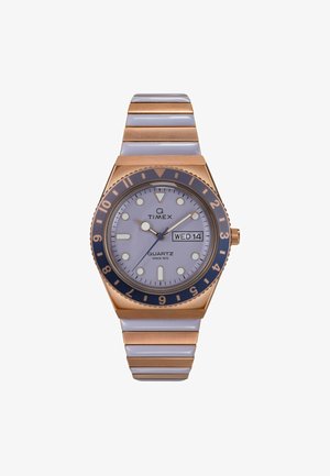 Timex Q kellaga, millel on ümmargune hõbedane nägu, roosa kuldne aktsent, sinine raam, integreeritud metallkäevõru ja kuupäeva näidik helesinisel dialil.