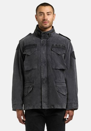 Mand iført en falmet sort militærinspireret jakke med flere forlommer og en høj krave, stående mod en ensfarvet baggrund.