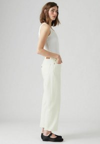 Witte, wijde jeans met voorzakken en een textuurafwerking; gecombineerd met een aansluitend wit hemdtopje en zwarte platte schoenen.
