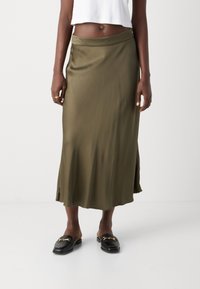 Vihreä oliviin sävyinen satiininen midi-hame, jossa on sileä rakenne, hienovarainen kiilto ja sivuaukot. Yhdistetty mustiin loafer-kenkiin, joissa on kultaiset yksityiskohdat.