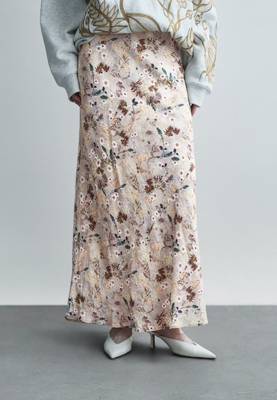 Jupe midi à motif floral en beige doux, ornée de broderies détaillées dans des tons de brun, vert et blanc. Texture lisse, associée à des talons blancs.