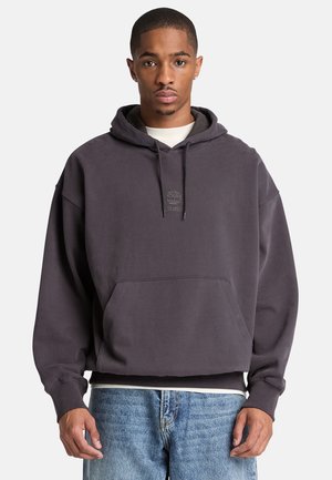 Mann trägt dunkelgrauen Timberland-Hoodie und hellblaue Jeans, steht mit neutralem Gesichtsausdruck vor einfachem Hintergrund.