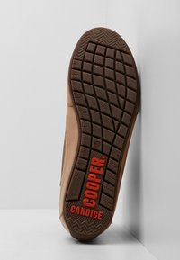 Semelle d’une chaussure marron avec un motif détaillé de la bande de roulement et un texte rouge "COOPER" et "CANDICE" en bas, appuyée contre un coin blanc.