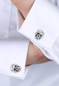 Silver skull cufflinks attached to a white dress shirt. The design features intricate facial details and a polished metallic finish.