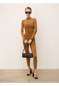 Rjava zgibana dolg rokav top, ujemajoče legice, ki prikazujejo teksturirano tkanino. Manekenka drži majhno črno torbico in nosi črne čevlje s blok peto.