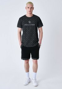 T-shirt noir avec un motif géométrique et le texte "PROJECT X PARIS" ; assorti à un short noir avec des finitions coordonnées et des baskets blanches.