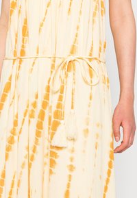 Żółto-biały sukienka w stylu tie-dye z paskiem w talii, zakończonym dwoma białymi frędzelkami. Lekka tkanina z pionowymi fałdami i zwiewnym krojem.