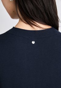 Ναυτικό μπλε πλεκτό πουλόβερ με ριμπ λαιμόκοψη και μια μικρή ασημένια λεπτομέρεια στην πλάτη. Λείο υφή, απλός σχεδιασμός, χωρίς ορατά μοτίβα.