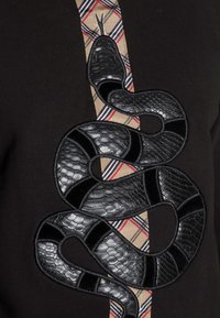 Prenda de punto negra con una prominente serpiente negra bordada y texturizada, rodeada por una tela a cuadros en tonos beige y rojo.