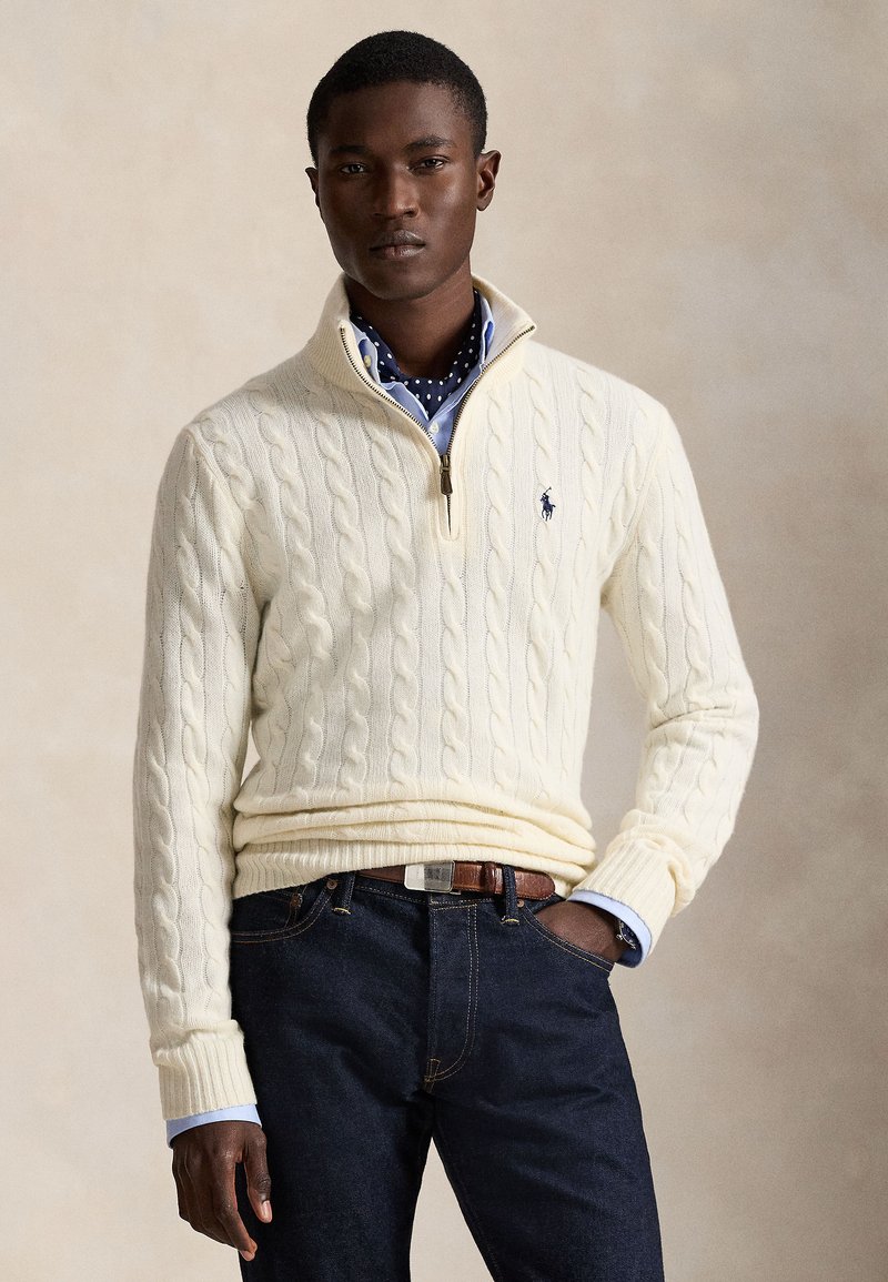 Uomo con maglione crema lavorato a trecce con mezza zip sopra una camicia blu con sciarpa a pois, jeans scuri e cintura marrone su sfondo neutro.