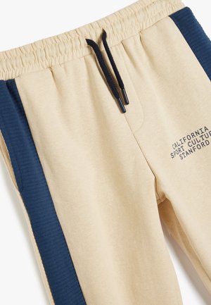Pantalones de chándal beige con paneles laterales azul oscuro, cinturilla elástica y cordón negro. Presenta texto impreso en la parte frontal. Textura de tela suave.