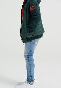 Grön puffjacka med dragkedja framtill, svarta detaljer, stickade ärmslut och ett märke på armen, parat med ljusblå skinny jeans och vita sneakers.