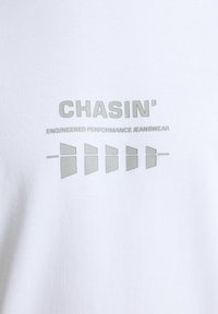 Camiseta blanca de algodón con una impresión mate gris que incluye la palabra "CHASIN'" y las palabras "ENGINEERED PERFORMANCE JEANSWEAR", además de un gráfico con cinco formas.