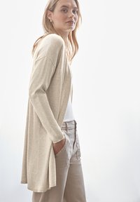 Lång beige kardiagan med avslappnad passform och ribbade muddar, bärs över en vit topp, i kombination med ljusbeige byxor. Slät textur.