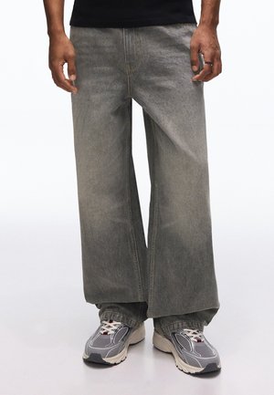 Personne portant un jean gris ample et des chaussures de sport grises, debout contre un fond blanc, les mains reposant le long du corps.