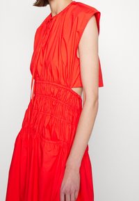 Ärmlös ljusröd klänning med rynkad midja, sidokut och knytning i halsen på en person med ljus hy.