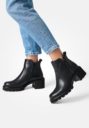 Schwarze Lederstiefeletten mit einem robusten Absatz und Gummisohle. Sie verfügen über einen seitlichen Reißverschluss und eine glatte Oberfläche, kombiniert mit hellblauen Jeans.
