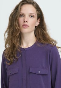 Giovane donna con lunghi capelli castani ondulati, indossa una camicia viola con bottoni e due tasche frontali, si trova di fronte con un'espressione neutra.