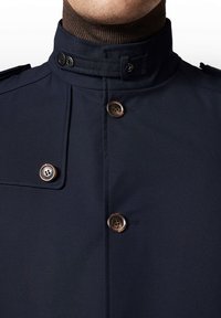 Granatowy płaszcz z wysokim kołnierzem, z dwoma ciemnymi guzikami i brązowym prążkowanym dekoltem. Gładki materiał o strukturalnym wzorze z epauletami na ramionach.