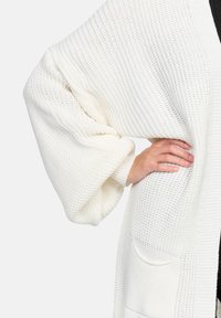 Vit stickad cardigan med breda ärmar, lös passform och en framficka. Strukturerad med en andningsbar öppen vävdesign.