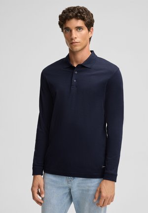 Marineblaues Poloshirt mit langen Ärmeln, klassischem Kragen, Knopfleiste mit drei Knöpfen und strukturierter Oberfläche; kombiniert mit hellblauen Jeans.