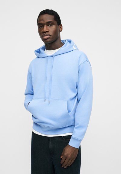 Homme portant un sweat à capuche bleu clair avec poche avant et cordons, associé à un pantalon noir, sur un fond gris clair uni.