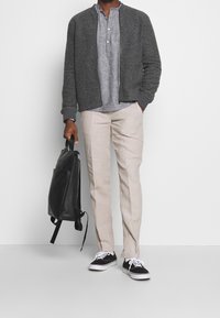 Harmaa bomber-takki, vaaleanharmaa henley-paita, beigehousut, musta reppu ja mustat tennarit valkoisilla pohjilla.