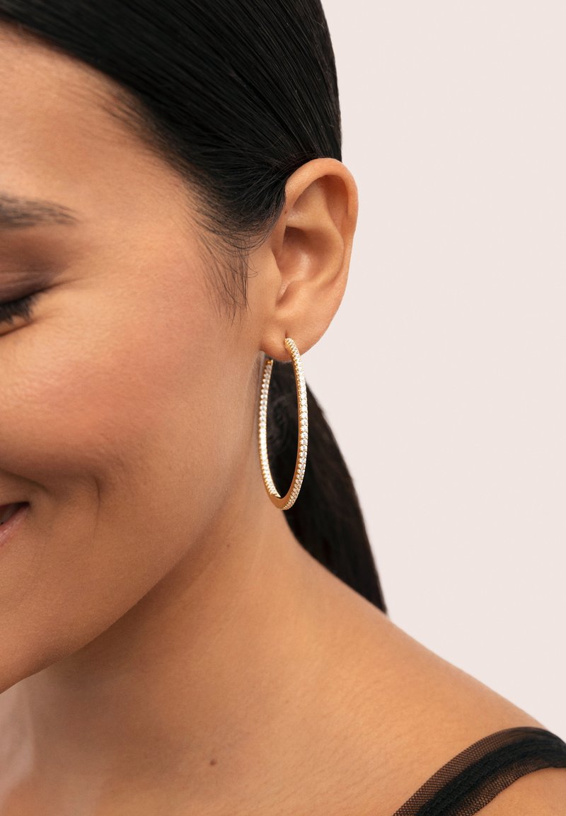 Pendientes de aro dorados con una superficie texturizada, con una línea de pequeñas piedras transparentes a lo largo del borde. Llevados por una persona con cabello oscuro.