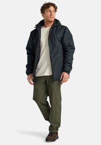 Hombre vestido con chaqueta con capucha negra, camisa beige, pantalones verde oliva y zapatos de senderismo marrones, de pie con una pierna cruzada y mirando hacia la derecha.